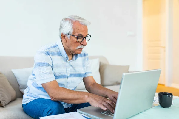 Mature businessman sit on sofa working on computer wireless device using e-banking application paying bills online, typing e-mail solve issues distantly, older generation and modern tech usage concept