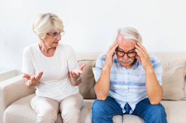 Elderly couple arguing. Senior man having headache, holding his head between hands. Mature woman frustrated with her husband. Marrige problems