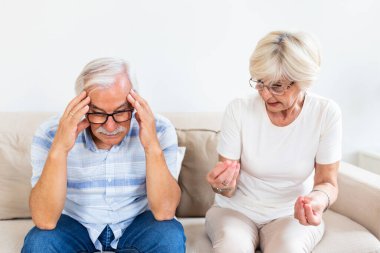Elderly couple arguing. Senior man having headache, holding his head between hands. Mature woman frustrated with her husband. Marrige problems