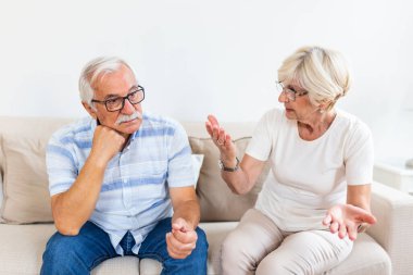 Senior couple having argument on the couch at home in the living room. Woman and man emotions gesturing with hands. Senior couple arguing