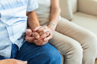 Close up of mature old husband and wife hold hands show love and care. Senior couple holding hands while sitting together at home. Elderly relationships, marriage concept