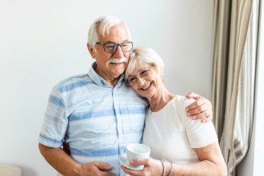 Happy senior couple standing in the living room. Looking each other smilling and holding cup of coffee or tea. Elderly husband hugging his lovely wife