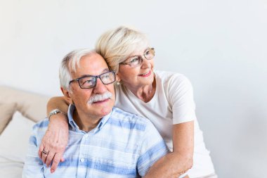 Portrait of overjoyed elderly husband and wife sit on couch hugging looking in distance. Happy mature spouses couple embrace cuddle relax together on sofa, enjoy time together
