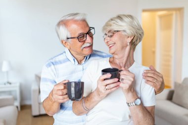 Happy senior couple standing in the living room. Looking each other smilling and holding cup of coffee or tea. Elderly husband hugging his lovely wife