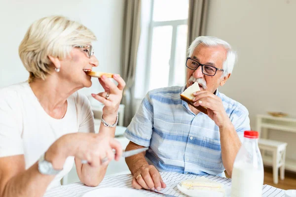 Senior couple eating breakfast at home. Holding piece of bread. Elderly couple enjoy in their time together. Feeling happy. Senior man eating sandwich