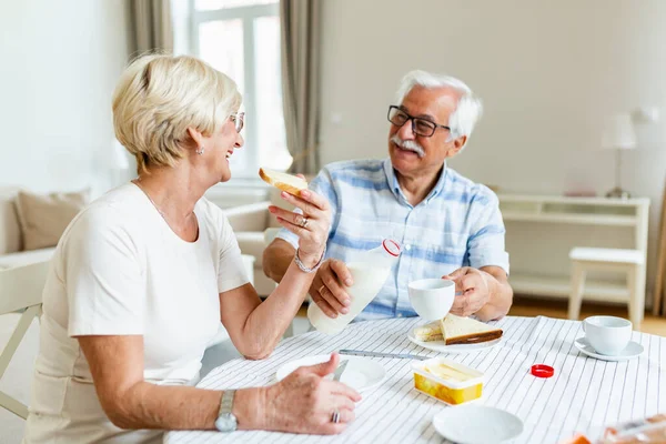 Elderly couple sitting at the table. Senior man pouring milk into a cup. Married couple having breakfast at home. Mature woman holding peice of bread