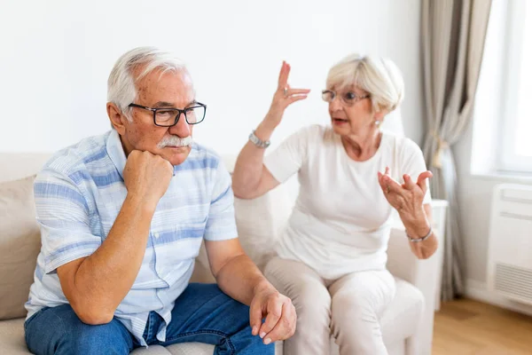 Senior couple having argument on the couch at home in the living room. Woman and man emotions gesturing with hands. Senior couple arguing