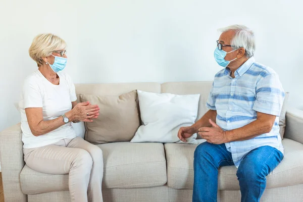 Two Elderly friends sitting in social distance wearing protective mask and talking on the sofa, preventing covid 19 coronavirus pandemic infection spread. Social distancing on sofa at home