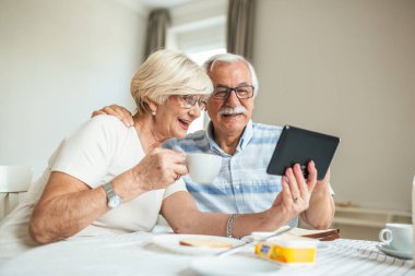 Senior man and his wife are using a digital tablet and communicating with their kids. Happy senior couple is having breakfast and having an online conversation with their family and sending greetings.
