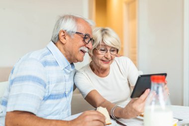 Senior couple smiling and looking at the same tablet hugged. Indoor, at home concept. Mature and retired man and woman using technology - lockdown and quarantine lifestyle