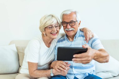 Couple of seniors smiling and looking at the same tablet hugged on the sofa - indoor, at home concept - caucasians mature and retired man and woman using technology - lockdown and quarantine lifestyle