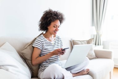 Pretty woman using phone and laptop at home texting. Happy young woman studying at home reading an sms or text message on her mobile phone with a smile as she lies on the sofa with a laptop