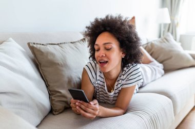 Smiling African American woman reading message on her mobile phone while sitting on sofa at home. Happy girl reading a message in a smart phone sitting on a couch at home