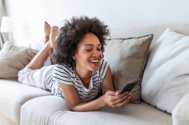 Pretty girl using her smartphone on couch at home in the living room. Young beautiful woman texting on her mobile phone while sitting at home in comfortable sofa