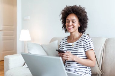 Happy young black woman using mobile phone while sitting a couch at home with laptop computer. Smiling woman sitting on sofa relaxing while browsing online shopping website.