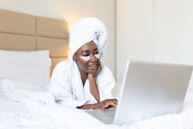Smiling african young woman with face cream under her eyes lying on bed in bathrobe and towel on the head using a laptop on the bed at home