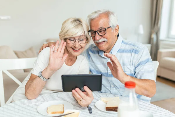 Happy older couple using computer tablet together at home, excited mature man and woman looking at mobile device screen, video call chatting online while having breakfast at home.