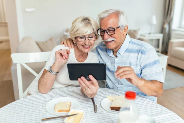 Happy mature couple using digital tablet and waving while having video call at home. Elderly couple making video call over touchpad while having breakfast at home.