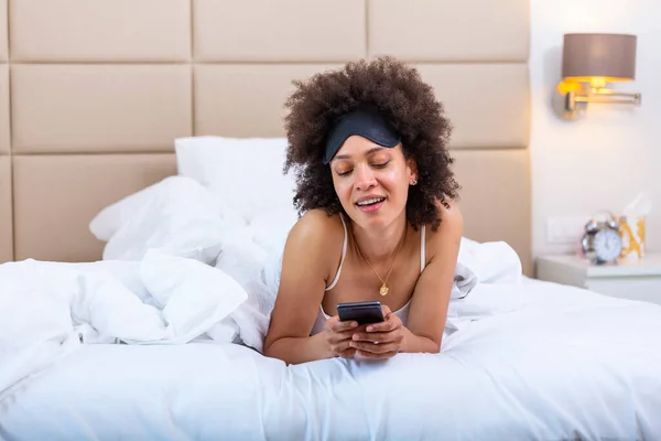 Beautiful woman with mobile phones lies in bed wearing sleeping mask. Happy young woman using cellphone at home. A woman is using her smartphone while she smiles lying on her bed.
