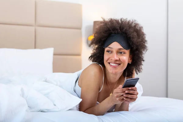 Happy excited woman texting on phone while wearing sleeping mask. Chatting with friend. Young woman lying on the bed in the bedroom and using mobile phone. Woman with mobile phone