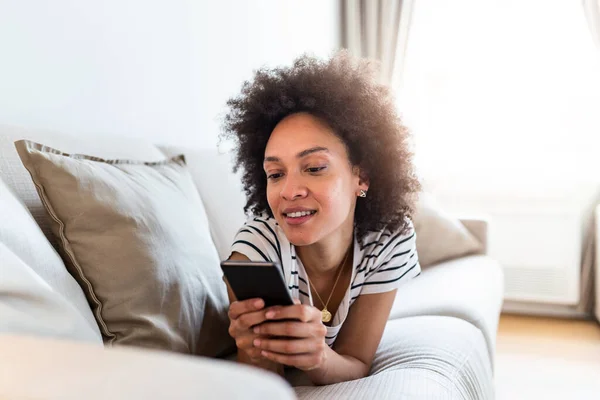 Happy young woman with mobile phone laying on sofa. Young happy woman lie down on bed and playing smart phone at home. Beautiful young black women using tablet computer