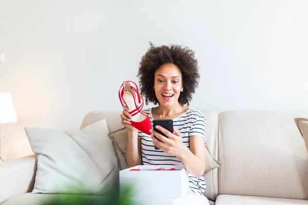 Smiling woman unboxing a postal parcel and taking a selfie with her new purchases using a smartphone. Showing shoes to her friends via video call