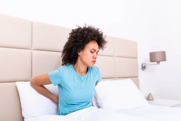 Hurt african American young woman sit in white bed wake up touch back suffering from painful backache, unwell black millennial girl awake from sleep having strong spinal muscular spasm strain