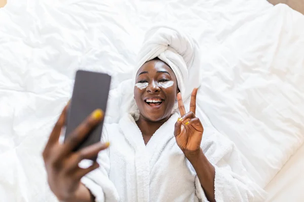 Smiling african woman lying on bed in bathrobe with mobile phone taking a selfie. African woman with eye mask relaxing on the bed after bath and looking at the phone camera taking a selfie