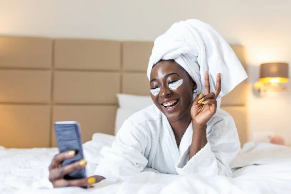 Smiling african woman lying on bed in bathrobe with mobile phone taking a selfie. African woman with eye mask relaxing on the bed after bath and looking at the phone camera taking a selfie