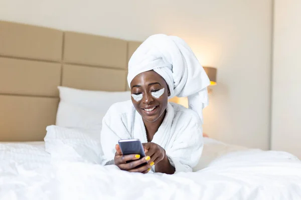 Attractive young black girl in bathrobe and with a towel on her head with eye mask is holding mobile phone,texting and smiling while sitting on a bed. Young African woman on bed