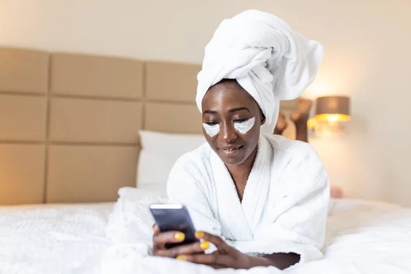 Attractive young black girl in bathrobe and with a towel on her head with eye mask is holding mobile phone,texting and smiling while sitting on a bed. Young African woman on bed