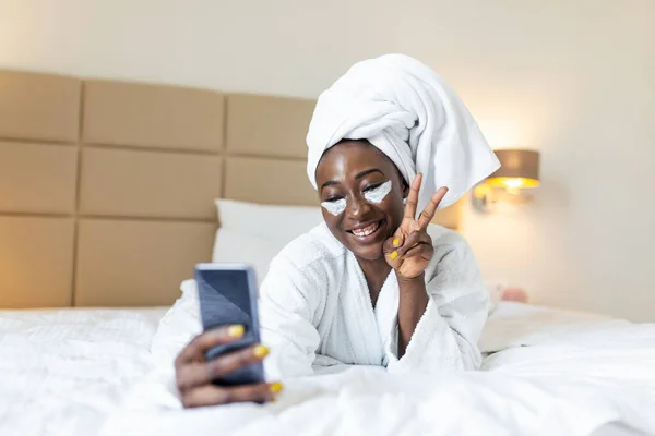 Smiling african woman lying on bed in bathrobe with mobile phone taking a selfie. African woman with eye mask relaxing on the bed after bath and looking at the phone camera taking a selfie