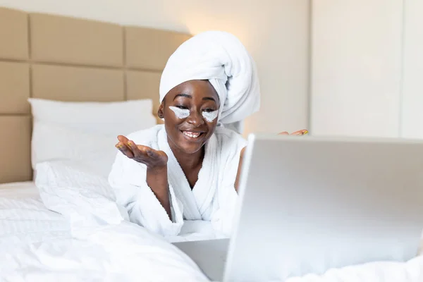 Beautiful young African woman in bathrobe resting on bed after shower video calling her friends on her laptop in the morning.Looking at laptop computer and talking to her friends via video call.