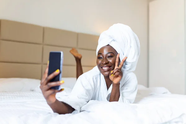 Smiling african woman lying on bed in bathrobe with mobile phone taking a selfie. African woman relaxing on the bed after bath and looking at the phone camera taking a selfie showing peace sign