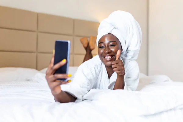 Smiling african woman lying on bed in bathrobe with mobile phone taking a selfie. African woman relaxing on the bed after bath and looking at the phone camera taking a selfie showing thumbs up