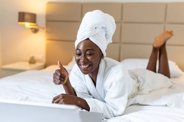 Beautiful young African woman in bathrobe resting on bed after shower while working on her laptop in the morning.Looking at laptop computer and talking to her friends via video call.