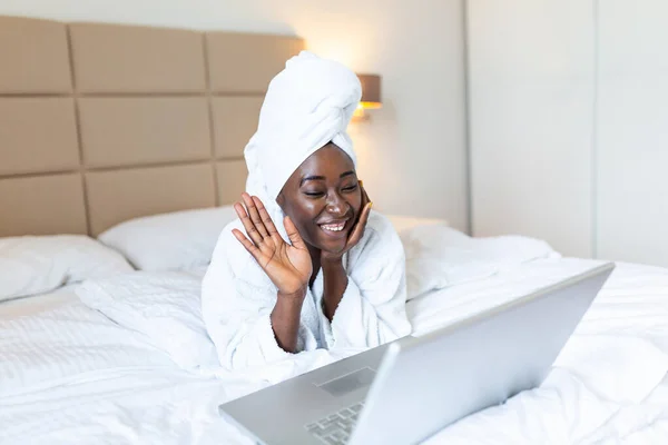 Smiling african woman lying on bed in bathrobe with laptop talking to her friends via video call. African woman relaxing on the bed after bath and looking at her laptop