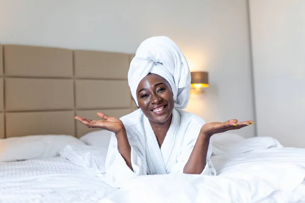 Young black woman in towel and bathrobe sitting on bed at home. beautiful African young woman wearing a white bathrobe, with a towel on her head in bed at home