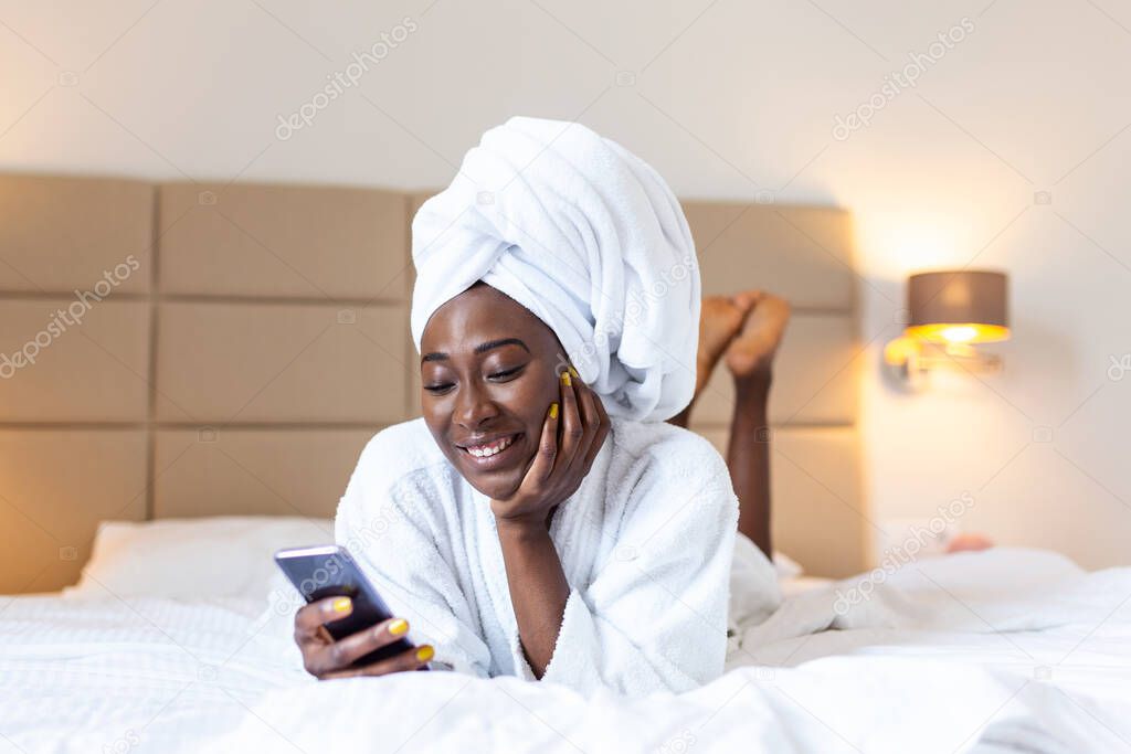 Beautiful young African woman in bathrobe resting on bed after shower at home. Young black woman wearing a towel and a white bathrobe holding mobile phone,texting and smiling