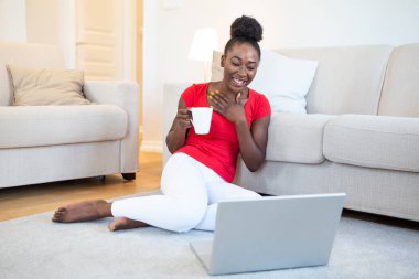 African american young woman having video call with friends or family. Holding cup of coffee or tea. sitting on the floor of living room. Beautiful woman feeling happy