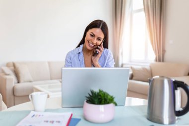 Smiling female multitask working on computer in office talking over phone, happy woman worker using laptop, laughing speaking with friend on cell or communicating with client online, work from home