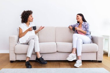 Two female friends in social distancing sitting on sofa in coronavirus pandemic time. Best friends having coffee together while separated by social distancing on sofa at home