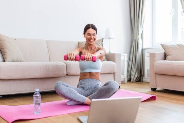 Young woman exercise with dumbbells at home and watching tutorial lesson on laptop computer. happy young female doing arm workout using dumbbells and looking at tutorial on her laptop