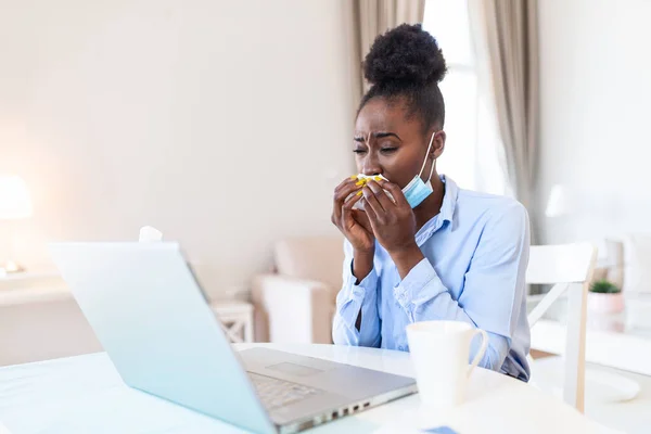 Young black businesswoman blowing her nose while working in her home office. Women are sneezing and are cold. She is in the office. Businesswoman wearing mask during COVID-19 pandemic.