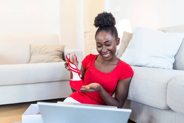 Smiling woman unboxing a postal parcel and showing shoes to her friends via video call. Young african American woman unpacking parcels with footwear, feeling happy with new online purchases