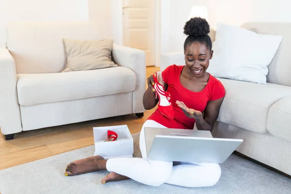 Smiling woman unboxing a postal parcel and showing shoes to her friends via video call. Young african American woman unpacking parcels with footwear, feeling happy with new online purchases