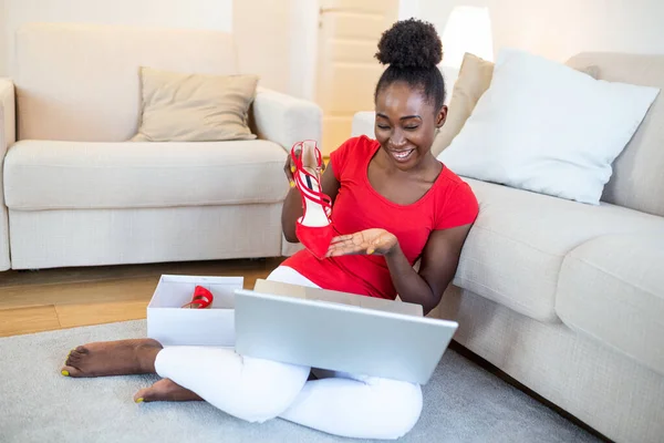 Smiling woman unboxing a postal parcel and showing shoes to her friends via video call. Young african American woman unpacking parcels with footwear, feeling happy with new online purchases