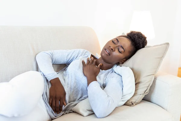 African beautiful woman sick with feeling pain in the chest while laying on the sofa .Hand holding chest after taking a medication.Concept of hard work without maintaining health.