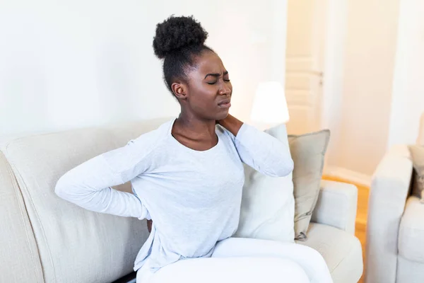 Young Black woman suffering from backache at home. Portrait of a young girl sitting on the couch at home with a headache and back pain. Beautiful woman Having Spinal Or Kidney Pain