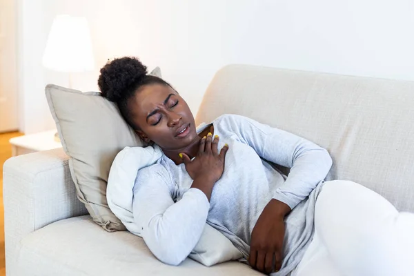 African beautiful woman sick with feeling pain in the chest while laying on the sofa .Hand holding chest after taking a medication.Concept of hard work without maintaining health.